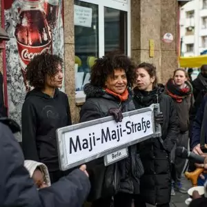 ISD, Dekoloniale, Jasmin Eding hält Straßenschild Maji-Maji-Str in der Hand umgeben von Leuten